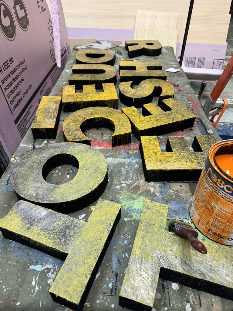 Large scenic letters beside an open Rosco paint can on a paint-covered shop table